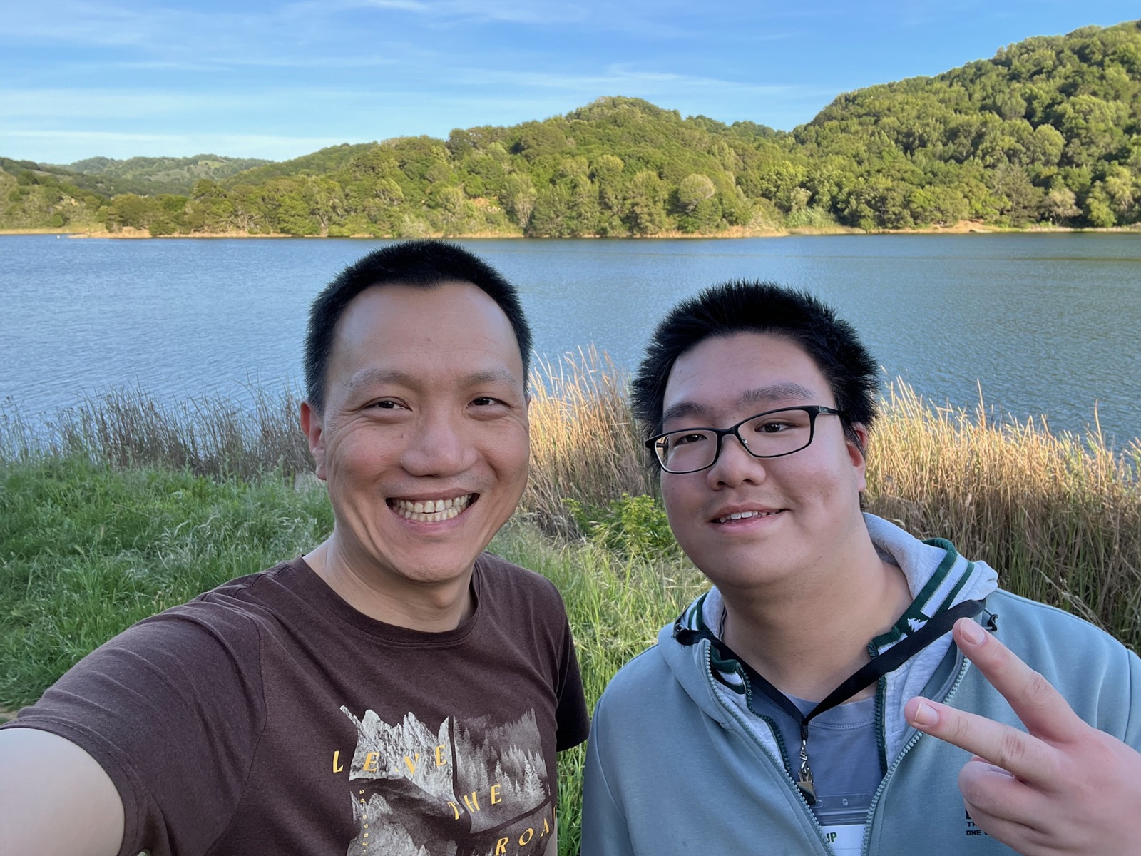 Two people smiling for a photo on a path beside a lake, with wooded hills behind them